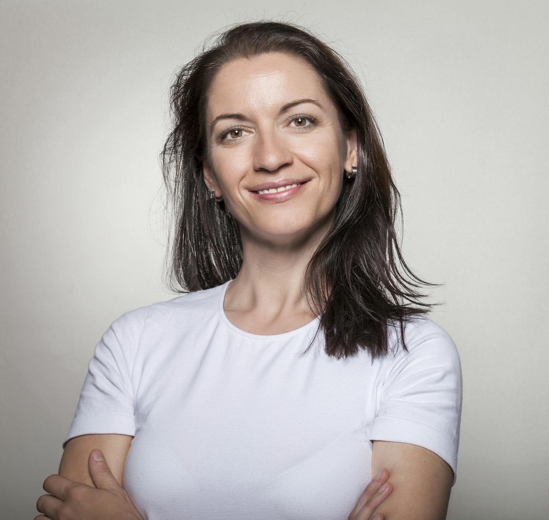 Portrait of a confident woman smiling with arms crossed, wearing a white t-shirt on a gray background.